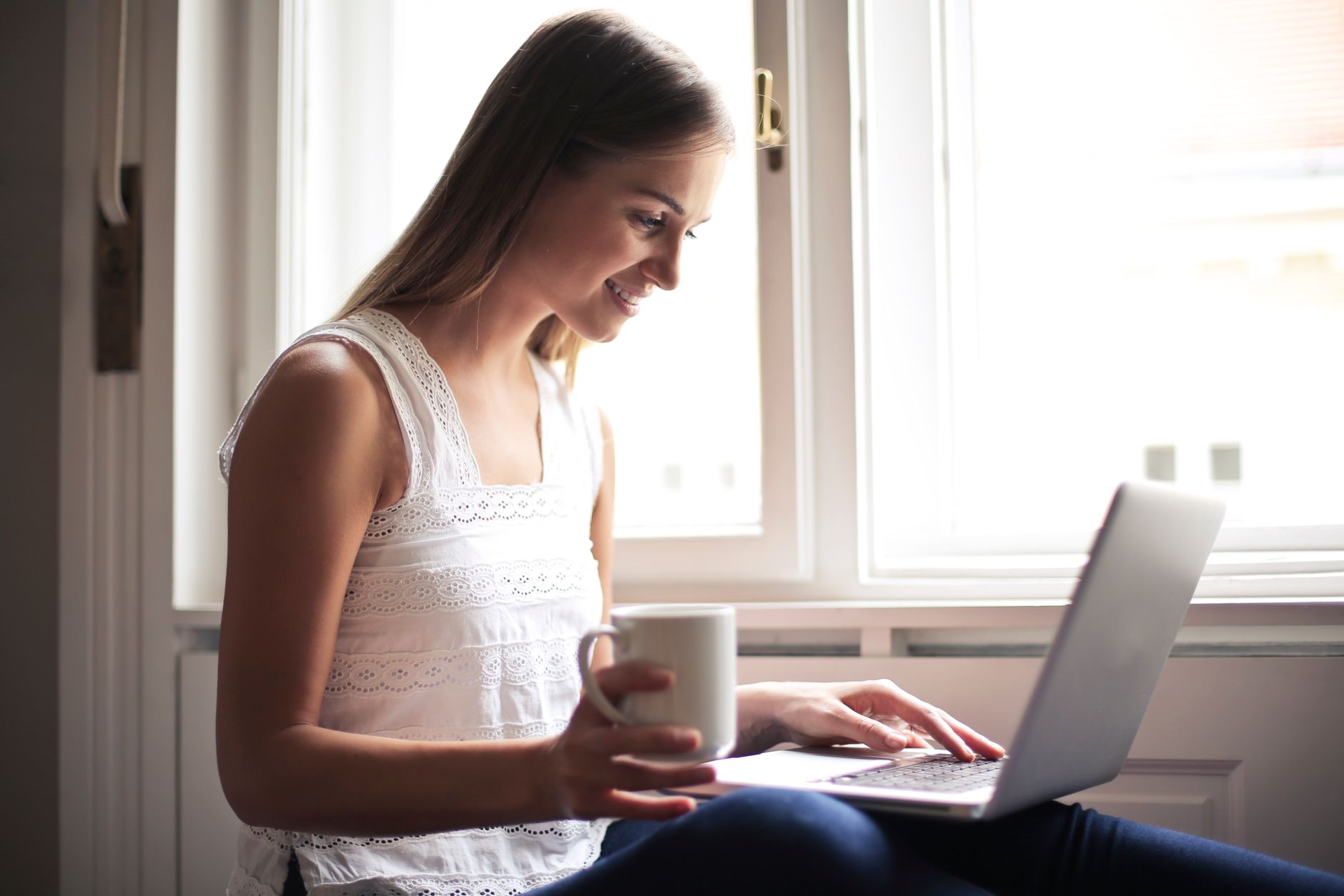Kvinna med kaffekopp kollar sin laptop hemma vid fönstret